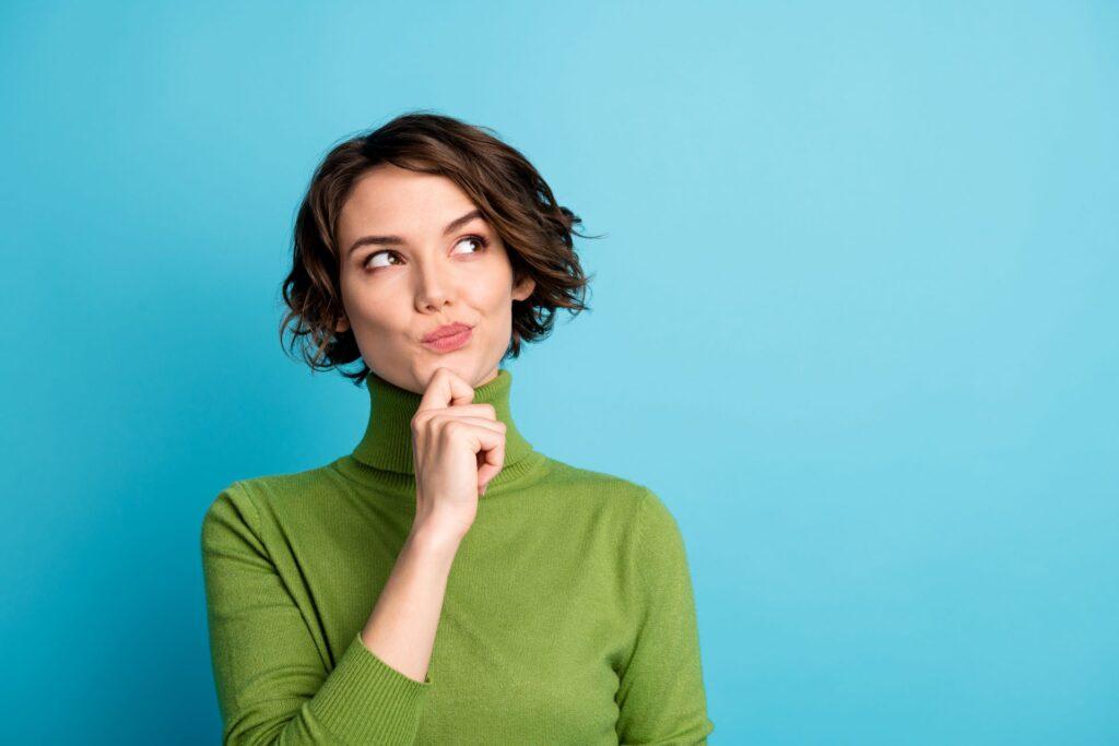 Junge Frau mit kurzen braunen Haaren, die nachdenklich schaut und ihren Finger an das Kinn legt. Sie trägt einen grünen Pullover und steht vor einem blauen Hintergrund.