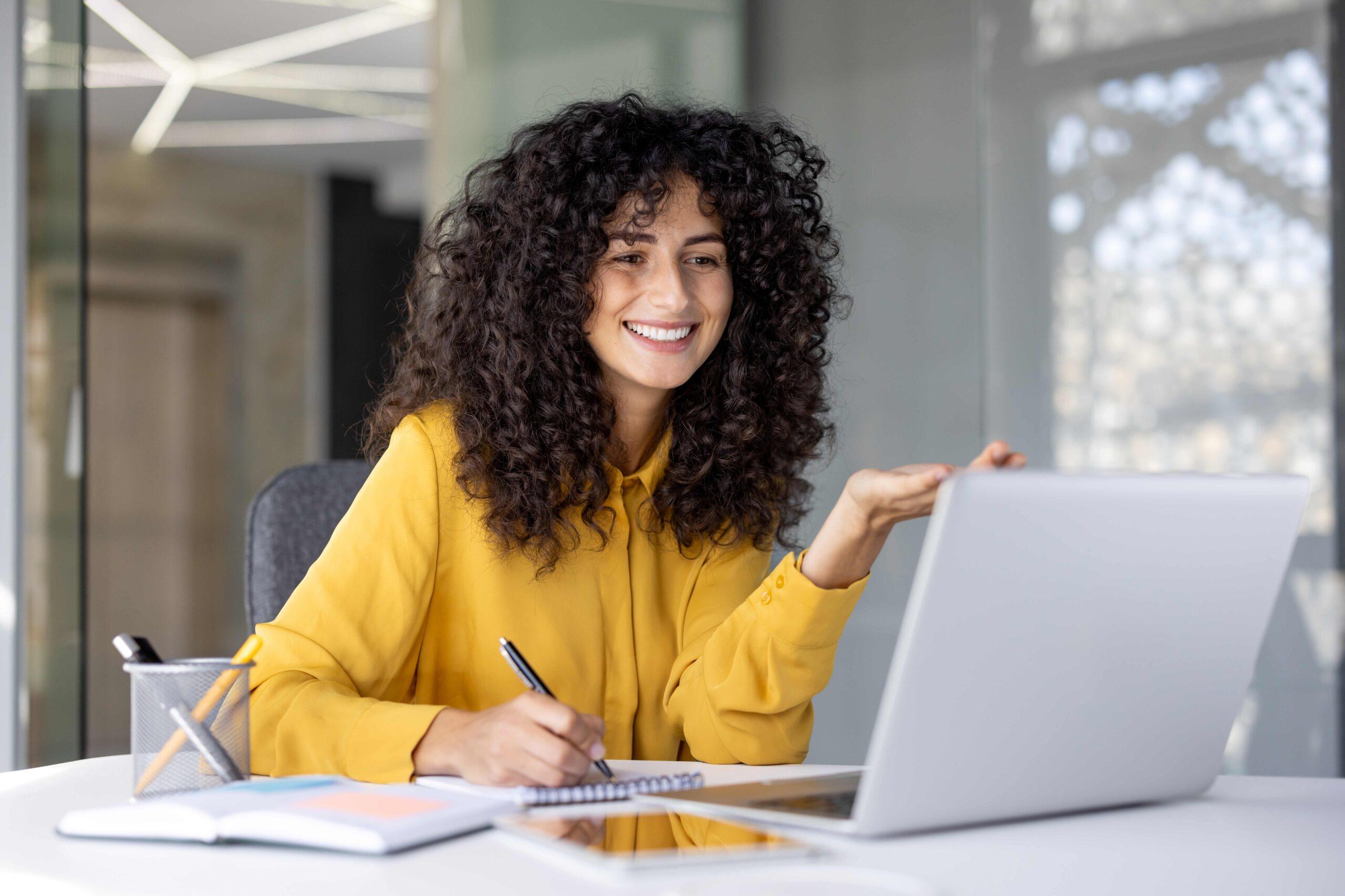 Eine lächelnde Frau in einem gelben Shirt nimmt an einem Online-Kurs teil, macht Notizen und gestikuliert vor ihrem Laptop.