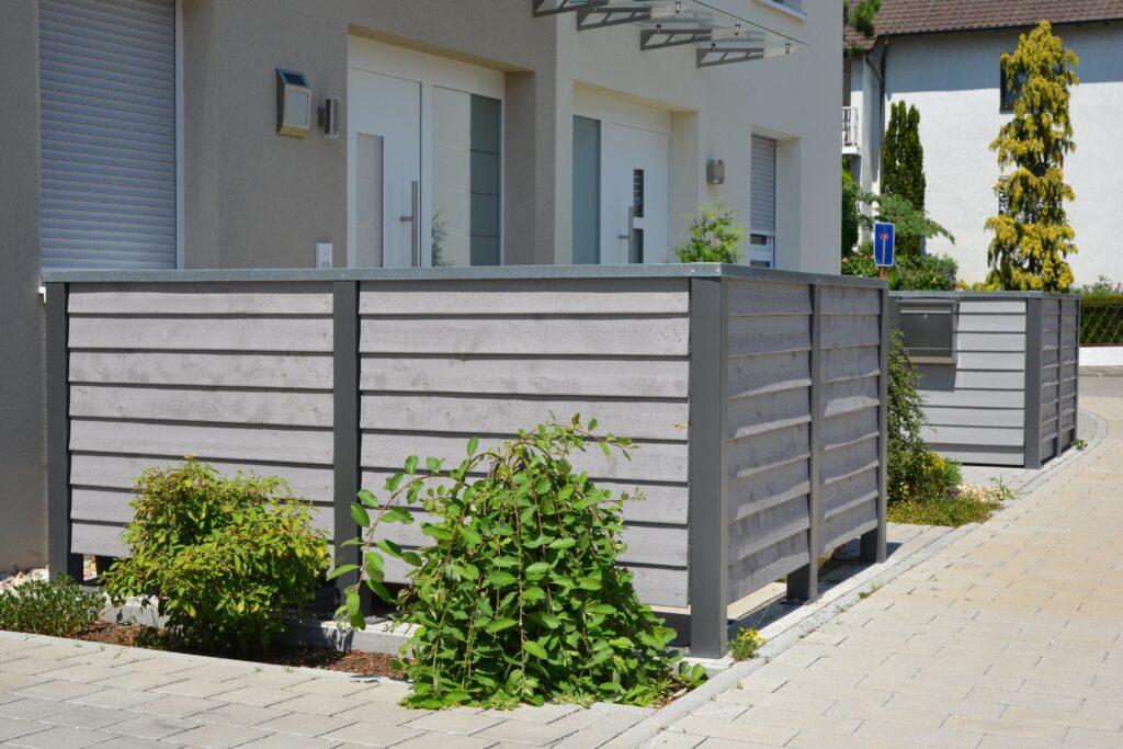 Moderne Mülltonnenverkleidung aus Holz vor einem Mehrfamilienhaus mit gepflegtem Vorgarten
